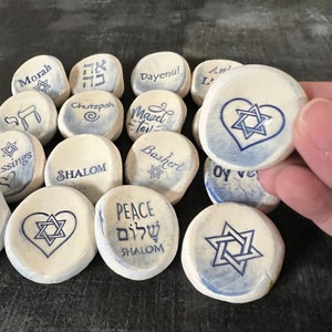 May include: A collection of white, oval-shaped stones with blue text and symbols. The stones feature phrases like "Shalom," "Peace," and "Mazel Tov," along with Star of David symbols. One stone is being held by a hand.