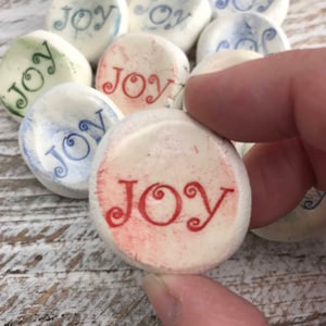 May include: A collection of small, smooth, off-white stones, each imprinted with the word "JOY" in various colors. The stones are held by a hand, with the word "JOY" in red ink on the front-most stone. Other stones have "JOY" in blue and green.