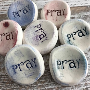 May include: A collection of seven ceramic stones, each with the word "PRAY" embossed in dark blue. The stones are in various pastel colors, including blue, pink, and white, with a distressed, textured finish.