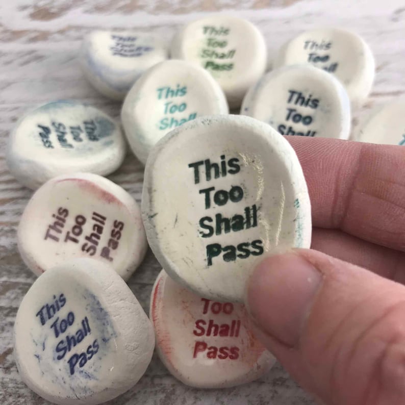 May include: A selection of smooth, white stones, each inscribed with the phrase "This Too Shall Pass" in different colours. The oval-shaped stones have a slightly textured surface, with text in black, blue, and red. A motivational item.