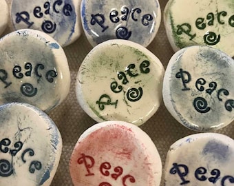 May include: A collection of small, round ceramic pieces with the word "Peace" inscribed in various colors. The pieces are primarily white with blue, green, and red lettering and decorative swirls. The overall aesthetic is handmade and artistic.