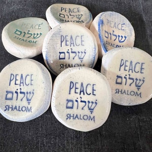 May include: A collection of seven white, round stones with the word "PEACE" and the Hebrew word "SHALOM" printed in blue. The stones have a slightly textured surface and vary in the intensity of the blue ink.