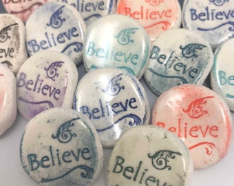 May include: A collection of smooth, oval-shaped stones with the word "Believe" inscribed in various colors. The stones feature decorative swirls and are in shades of blue, red, and purple. The overall theme is inspirational.