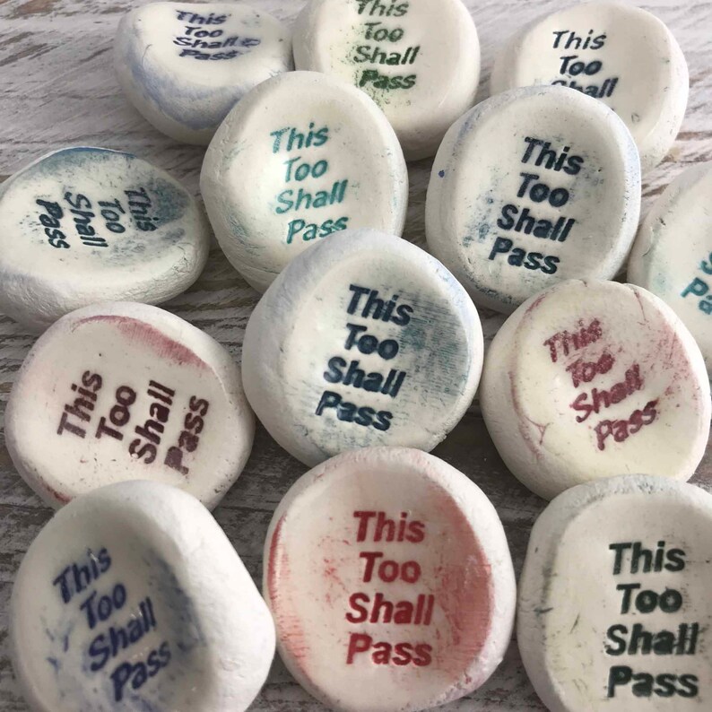 May include: A group of smooth, white stones, each inscribed with the words "This Too Shall Pass" in different colours. The text is in a plain font. The stones are displayed on a light-toned surface.