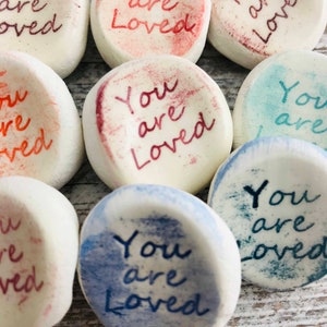 May include: Close-up of several white, rounded stones with the words "You are Loved" in various colors. The text is in a handwritten style, with red, blue, and teal hues. The stones have a slightly textured appearance.