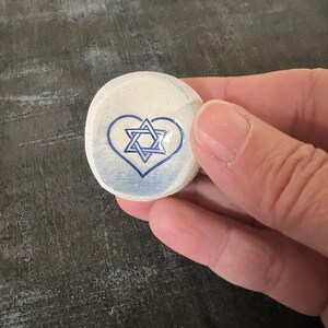 May include: A small, round, off-white stone with a blue heart and Star of David design. The stone is held in a hand against a dark gray background. The heart and star are painted in a simple, clean style.