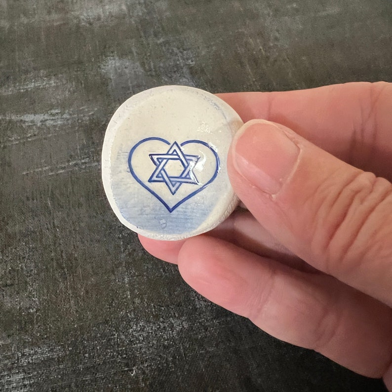 May include: A small, round, off-white stone with a blue heart and Star of David design. The stone is held in a hand against a dark gray background. The heart and star are painted in a simple, clean style.