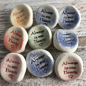 May include: Nine smooth, off-white stones with the inscription "Always in our Hearts" in various colors, including orange, blue, and burgundy. Each stone features a small heart symbol. The stones are arranged on a light-colored, textured surface.