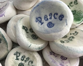 May include: A collection of white, smooth stones with the word "Peace" inscribed in various colors, including blue, green, and purple. Each stone has a unique design, with a spiral symbol. The stones are ideal for meditation or as decorative items.