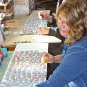 May include: A person smiling while working on a craft project. The project appears to be a mosaic or similar art form, with small, colorful pieces arranged on a surface. Various tools and materials are visible.