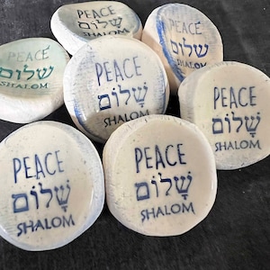 May include: A collection of white, oval-shaped stones with the word "PEACE" in English and Hebrew, along with the word "SHALOM". The text is printed in blue ink. The stones vary slightly in color and texture.