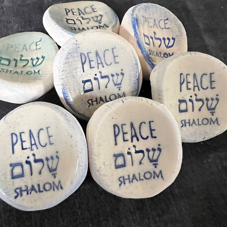 May include: A collection of white, oval-shaped stones with the word "PEACE" in English and Hebrew, along with the word "SHALOM". The text is printed in blue ink. The stones vary slightly in color and texture.