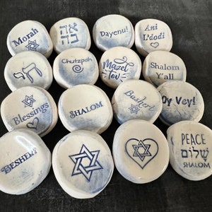 May include: A collection of white, smooth stones with blue text and Star of David designs. Each stone features a different word or phrase, including "Morah," "Dayenu!," "Blessings," and "Shalom." The stones are arranged on a dark surface.