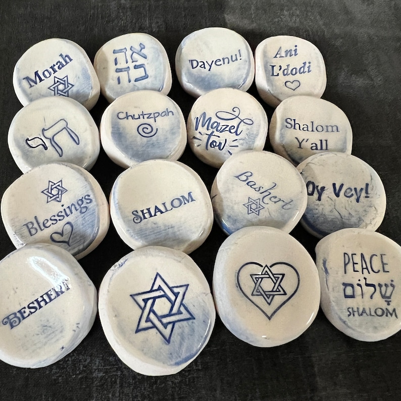 May include: A collection of white, smooth stones with blue text and Star of David designs. Each stone features a different word or phrase, including "Morah," "Dayenu!," "Blessings," and "Shalom." The stones are arranged on a dark surface.