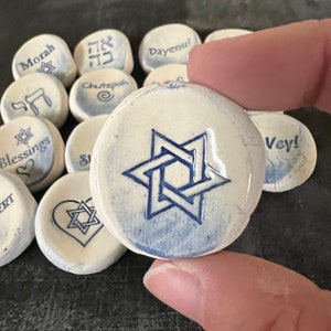 May include: A collection of white ceramic stones with blue text and designs. One stone features a blue Star of David. Other stones display words like "Blessings" and "Morah". The stones are round and appear handmade.