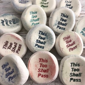 May include: A selection of smooth, white stones, each inscribed with the phrase "This Too Shall Pass" in different colours. The text is in a classic serif font, and the stones have a slightly aged appearance.