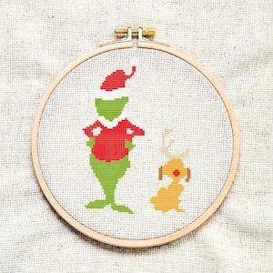 Puede incluir: Un bastidor de bordado de punto de cruz con un personaje verde con gorro y suéter rojos de Papá Noel, con un perro amarillo. El diseño está sobre tela blanca, en un aro de madera, y es una manualidad con temática navideña.