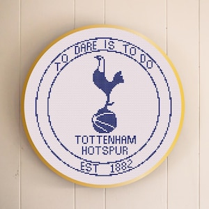 May include: A round, cross-stitched emblem featuring the Tottenham Hotspur logo. The design includes a navy blue rooster on a ball, surrounded by text that reads "TO DARE IS TO DO" and "TOTTENHAM HOTSPUR EST 1882". The emblem has a gold-colored border.