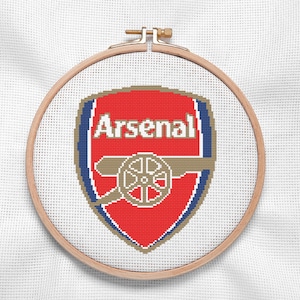 Könnte beinhalten: Kreuzstich-Stickerei des Logos des Fußballvereins Arsenal. Das Design zeigt ein rotes Schild mit dem Wort "Arsenal", eine Kanone und blaue und goldene Akzente. Die Stickerei befindet sich in einem hölzernen Stickrahmen.
