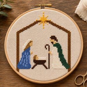 May include: A circular wooden embroidery hoop containing a cross-stitch pattern of a nativity scene. The design features a brown stable outline, a yellow star, and three figures in blue, white, and green garments surrounding a small manger.