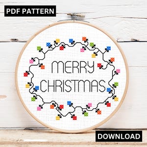 May include: A cross-stitch pattern in a wooden hoop features the words "MERRY CHRISTMAS" in black. The text is surrounded by a garland of colorful lights. The words "PDF PATTERN" and "DOWNLOAD" are in black boxes.