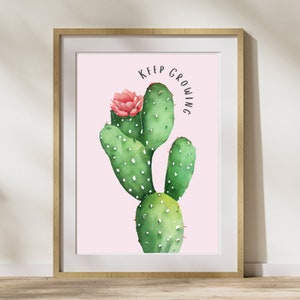 May include: A framed wall art print featuring a watercolor illustration of a green cactus with white spots and a single pink flower. The text KEEP GROWING is printed in black above the cactus against a solid light pink background.