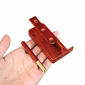 May include: A reddish-brown wooden rectangular case with a sliding lid featuring three circular indentations, accompanied by a matching wooden stick with a metallic gold-colored tip, presented as a two-piece set for storage or utility purposes.