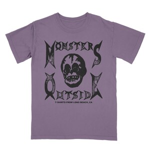 Może przedstawiać: Fioletowy T-shirt z czarnym nadrukiem graficznym czaszki z tekstem "Monsters Outside" i "T-shirts from Long Beach, CA".