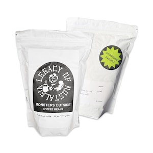 May include: Two white paper bags with black text and graphics. One bag has a black and white skull graphic with the text "LEGACY OF NOSTALGIA" and "MONSTERS OUTSIDE COFFEE BEANS". The other bag has a yellow starburst with the text "SURPRISE INSIDE!"