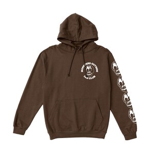 May include: A brown hoodie with a front pocket and a hood. The hoodie has a white graphic on the chest that says "MONSTERS OUTSIDE FAN CLUB" and white skull graphics down the left sleeve.