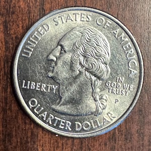 May include: A United States quarter dollar coin, featuring a profile of George Washington. The coin is silver in color, with the words "Liberty," "Quarter Dollar," and "In God We Trust" visible.