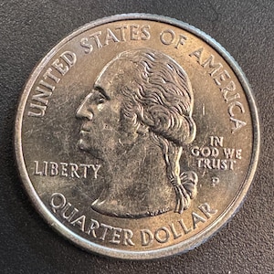 Może przedstawiać: Amerykańska moneta ćwierćdolarowa z profilem George'a Washingtona. Srebrna moneta zawiera napisy "Liberty", "Quarter Dollar", "United States of America" i "In God We Trust".