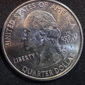 May include: A United States quarter coin with a portrait of George Washington. The coin is silver in color and features the words "Liberty", "Quarter Dollar", "United States of America", and "In God We Trust".