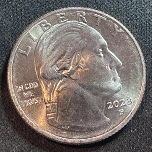 Può includere: Una moneta da venticinque centesimi di dollaro statunitense, con un profilo di George Washington, la parola "LIBERTY" in alto e l'iscrizione "IN GOD WE TRUST". L'anno 2023 è visibile, insieme alla lettera "P".