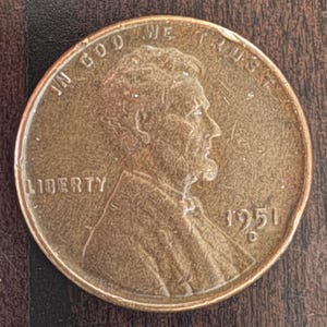Puede incluir: Una moneda de un centavo estadounidense de 1951 de color cobre. La moneda presenta un perfil de Abraham Lincoln, la palabra "LIBERTY" y la frase "IN GOD WE TRUST". También es visible el año "1951".