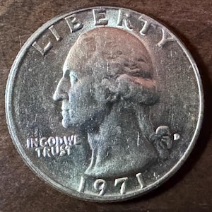 Może przedstawiać: Zbliżenie monety ćwierćdolarowej Stanów Zjednoczonych z 1971 roku. Moneta przedstawia profil George'a Washingtona z napisami "LIBERTY" i "IN GOD WE TRUST". Rok "1971" jest widoczny na dole.