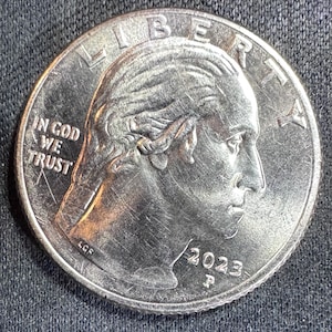 May include: A silver quarter coin with the profile of a person on the obverse. The coin features the inscriptions "LIBERTY" at the top, "IN GOD WE TRUST" on the left, and the year "2023" at the bottom. The coin is on a dark surface.