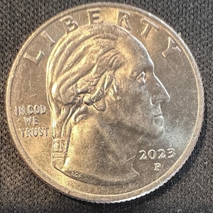 Può includere: Una moneta da venticinque centesimi di dollaro statunitense, con un profilo di George Washington. La moneta presenta l'iscrizione "LIBERTY" in alto e "IN GOD WE TRUST" in basso. L'anno "2023" è visibile, insieme alla lettera "P".