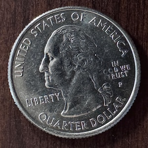Może przedstawiać: Srebrna moneta ćwierćdolarowa Stanów Zjednoczonych. Moneta przedstawia profil George'a Washingtona, napisy "LIBERTY", "QUARTER DOLLAR" i "UNITED STATES OF AMERICA". Widoczny jest również napis "IN GOD WE TRUST".