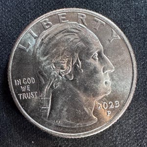 Può includere: Una moneta d'argento da un quarto di dollaro con il profilo di una persona rivolta verso destra. La moneta presenta le scritte "LIBERTY" e "IN GOD WE TRUST". L'anno "2023" è visibile.