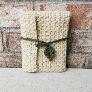 May include: A crocheted journal cover in a cream color, secured with a green crocheted tie and leaf accent. The cover is rectangular and appears to be handmade, with a textured stitch pattern. The background is a brick wall.