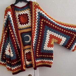 May include: A crocheted cardigan with a geometric pattern in shades of red, orange, yellow, blue, and white. The cardigan has a loose fit and long sleeves.