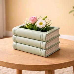 May include: A light green book-shaped planter, filled with a pink rose and a white daisy, sits on a round wooden table. The planter is designed to look like a stack of books. A decorative item for the home.