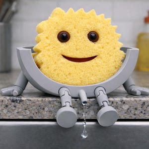 May include: A yellow sponge with a smiling face sits in a grey holder with legs. The holder is on a countertop, and a drop of water is falling from the bottom. Kitchen and cleaning supplies.