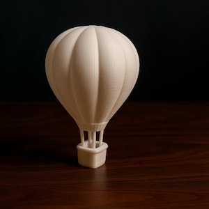 Può includere: Un piccolo modello di mongolfiera bianco sporco. Il pallone presenta dettagli a coste verticali ed è sostenuto da un cestino. Il modello è ambientato su uno sfondo scuro su una superficie di legno. Un oggetto decorativo.