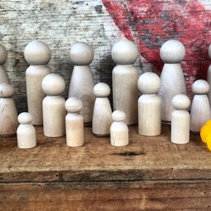 May include: A collection of wooden peg dolls of varying sizes, arranged in a row on a wooden surface. The dolls are all unpainted and have a natural wood finish. There are two orange flowers on either side of the dolls.