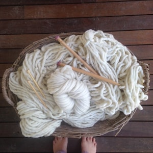 May include: A large basket filled with white yarn and two wooden knitting needles. The yarn is arranged in a loose, fluffy pile.