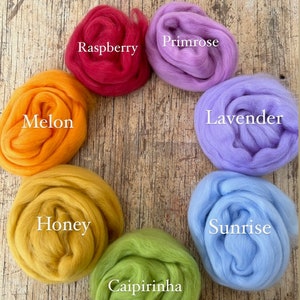 Puede incluir: Un arcoíris suave de vellón de lana en tonos de rojo, naranja, amarillo, verde, azul y morado. Los colores están etiquetados con nombres como Raspberry, Melon, Honey, Caipirinha, Sunrise, Lavender y Primrose.