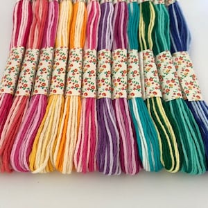 May include: A collection of colorful embroidery floss skeins. The floss is arranged in a row, showcasing a variety of colors including pinks, yellows, oranges, greens, and blues. Each skein is wrapped with a floral patterned paper.