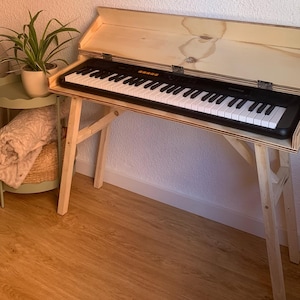 Può includere: Una tastiera in bianco e nero è all'interno di un supporto per pianoforte in legno con coperchio incernierato. Il supporto ha una finitura in legno naturale e gambe angolate. Un'opera d'arte bianca strutturata è appesa al muro sopra la tastiera.